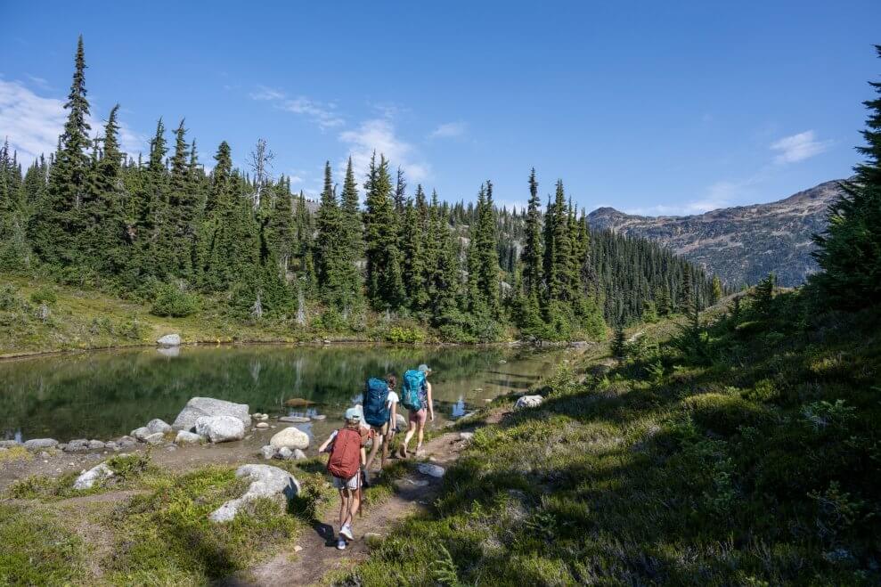A mom and her two kids wear backpacks as they hike along a lake in the mountains. Hiking games are a great way to add some fun to hikes with kids.