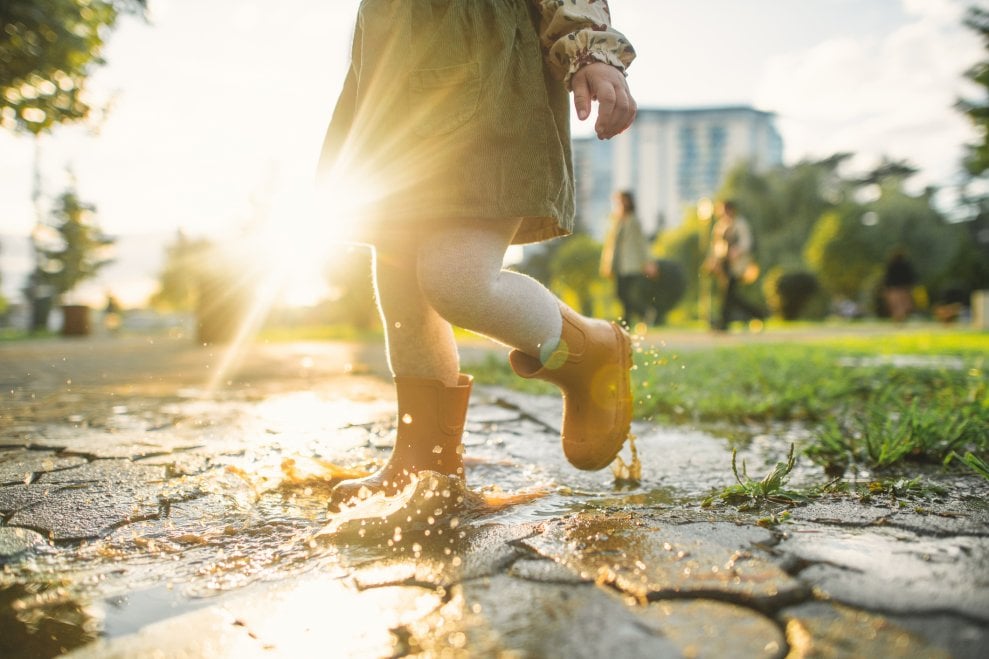 A girl wearing a dress and yellow rubber boots splashes in a puddle on a sunny day.