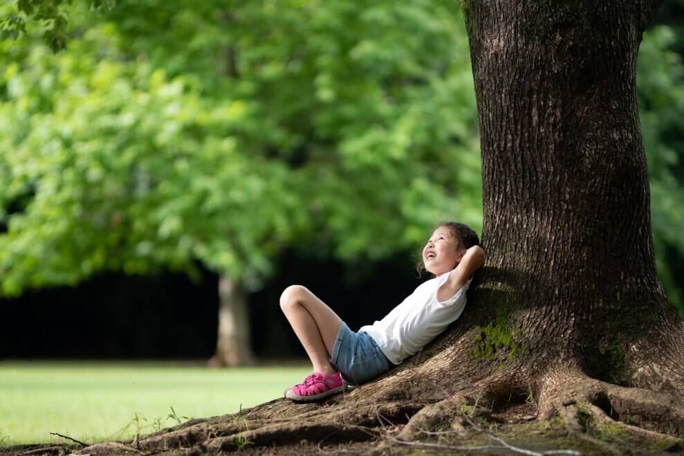 A girl sits under a tree, smiling up at the tree cover.