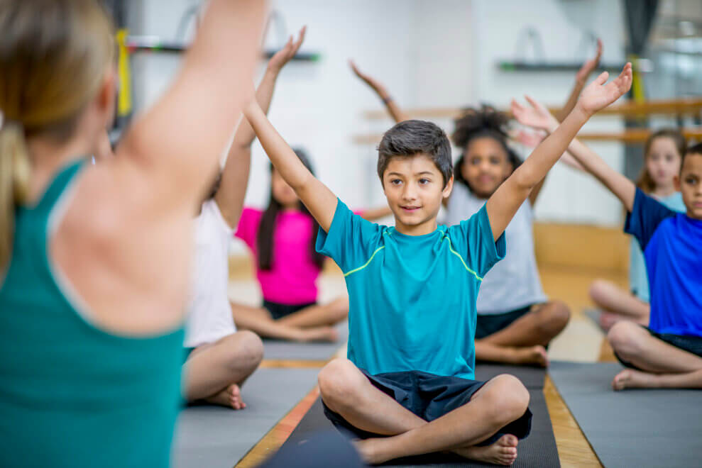 In a children's yoga class, kids sit on their mats in front of the instructor and reach their arms over their heads.