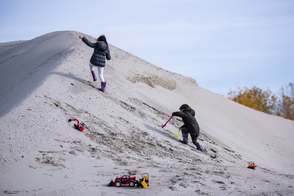 Two children play on a sand dune with toy shovels and trucks. They're both climbing up a sandy hill. 