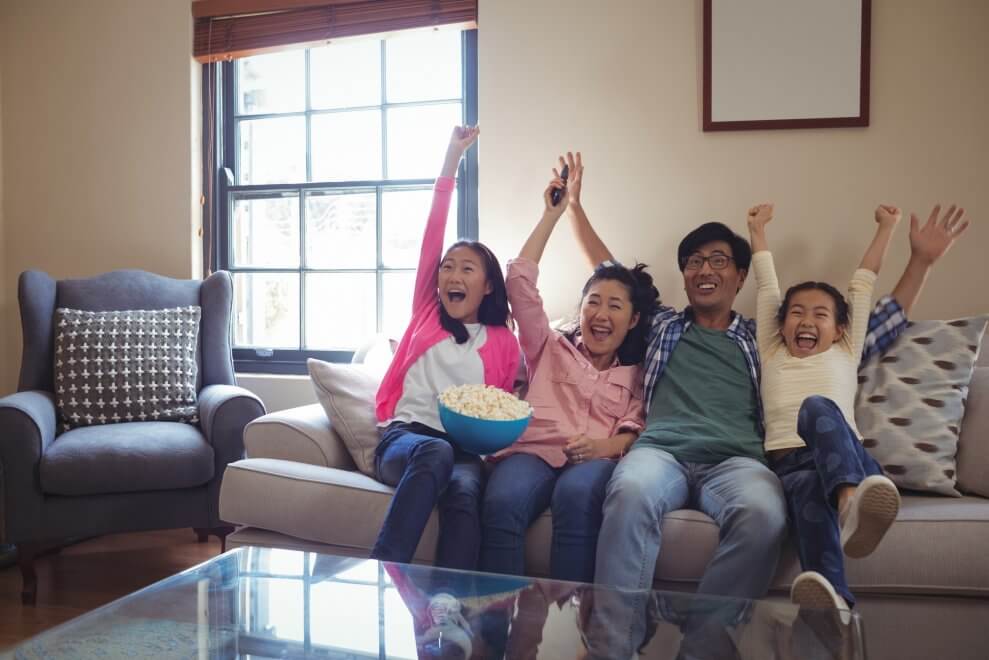 A father, mother, and their two daughters cheer as they watch TV together in their living room.