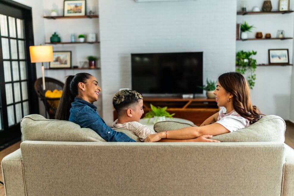 Two moms and their preteen son sit on the couch in the living room, talking. The TV behind them is turned off.