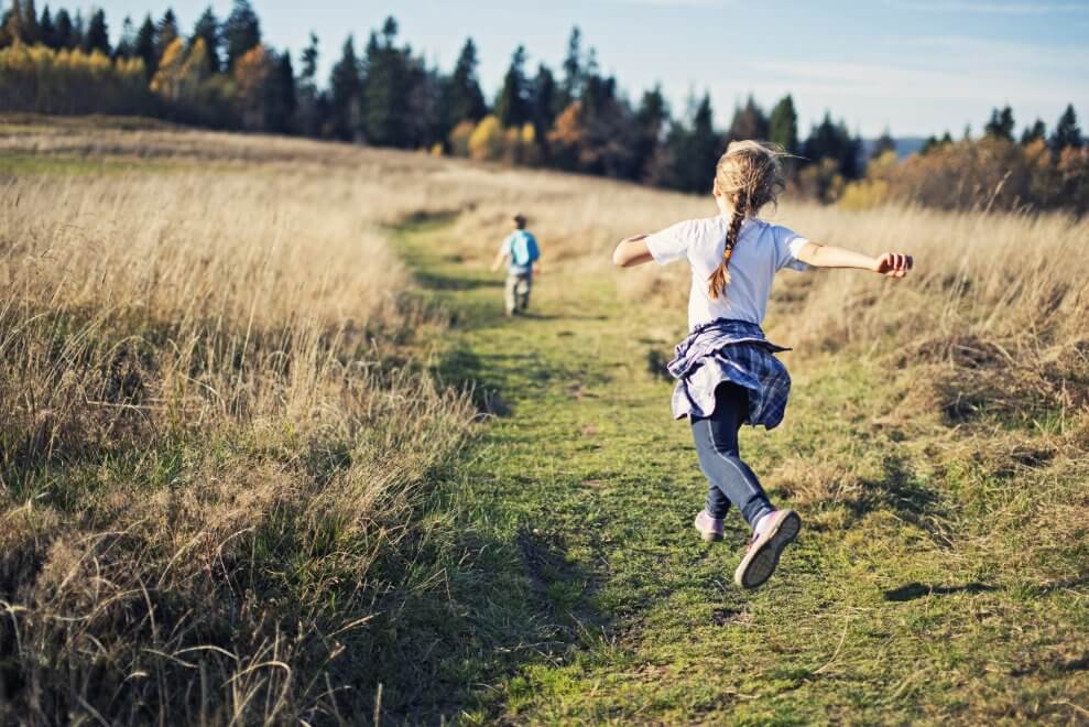 Two children happily run in a field on a fall day, with their backs to the camera.