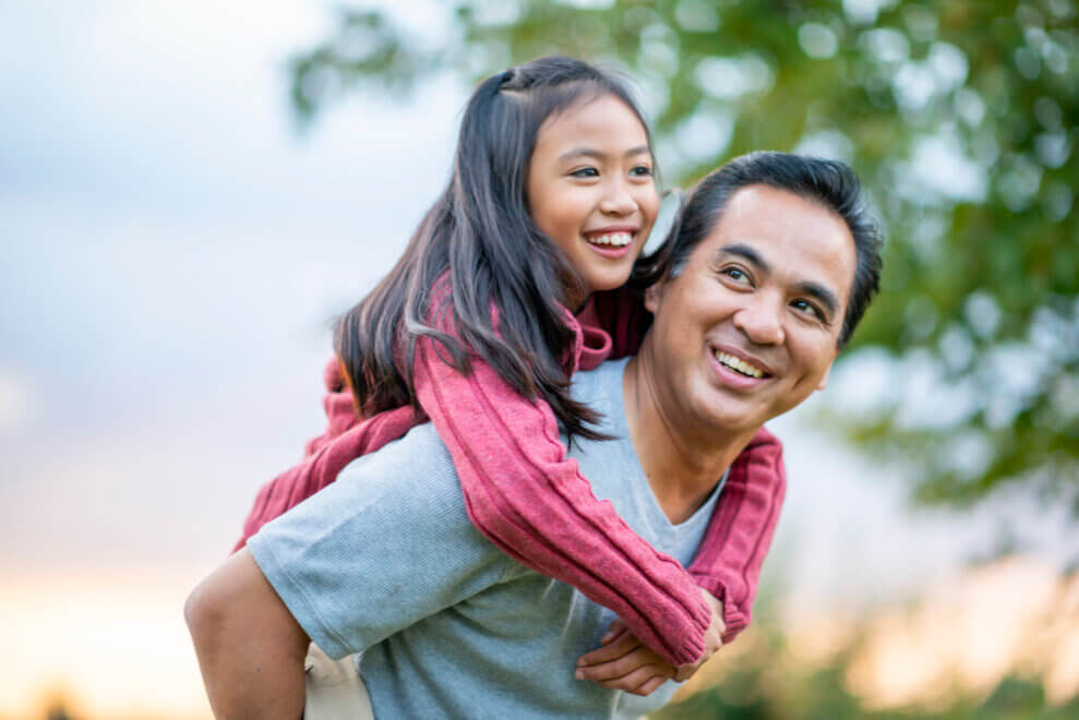 A father gives his daughter a piggy-back ride outside. They're both smiling.