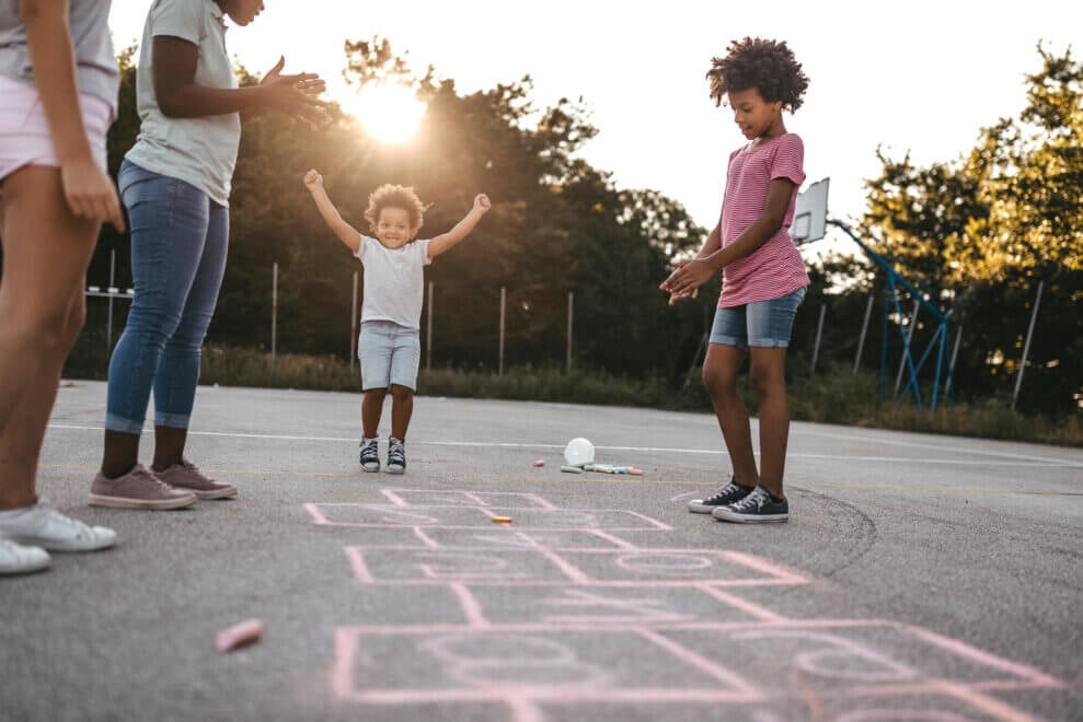 Family plays hopscotch outdoors, with one child cheering with her arms above her head