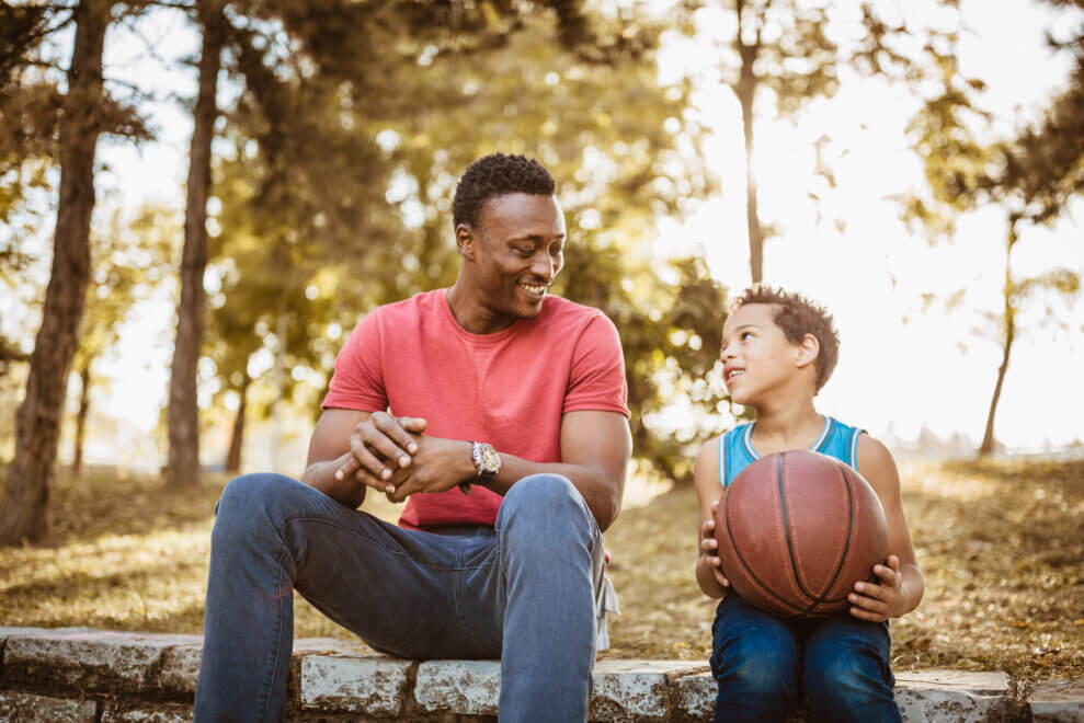 A father and his son sit outside in a park, talking. The son holds a basketball in his arms.