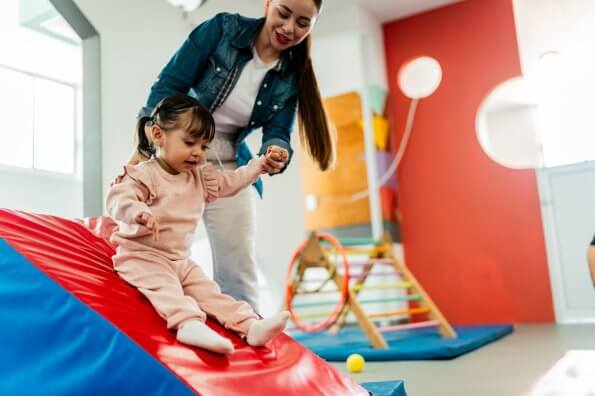 An early childhood educator helps a toddler slide down a foam slide.