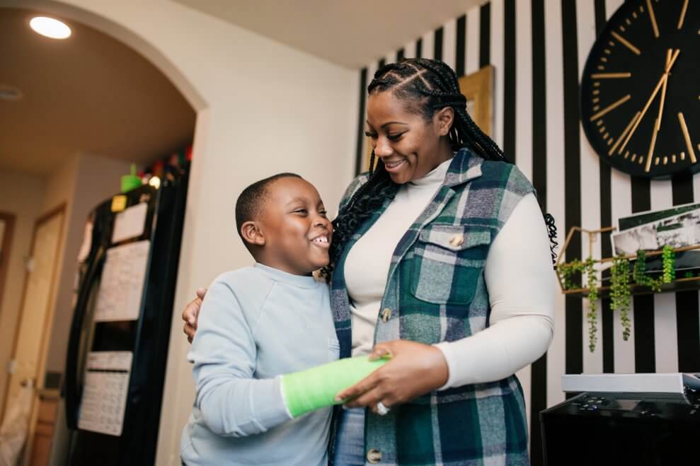 A boy wears a green cast on his wrist as he and his mom smile and hug in their living room.
