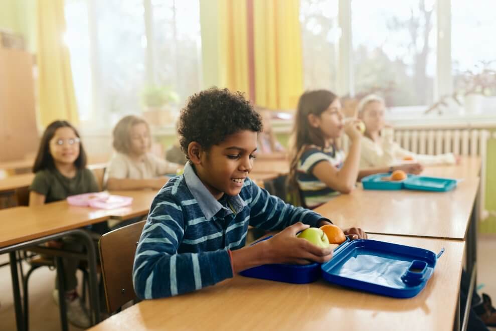 A boy sits at his desk at school and takes healthy snacks out of his lunch box.
