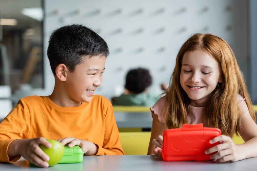 Two elementary students sit at their desk and take snacks out of their lunch boxes.