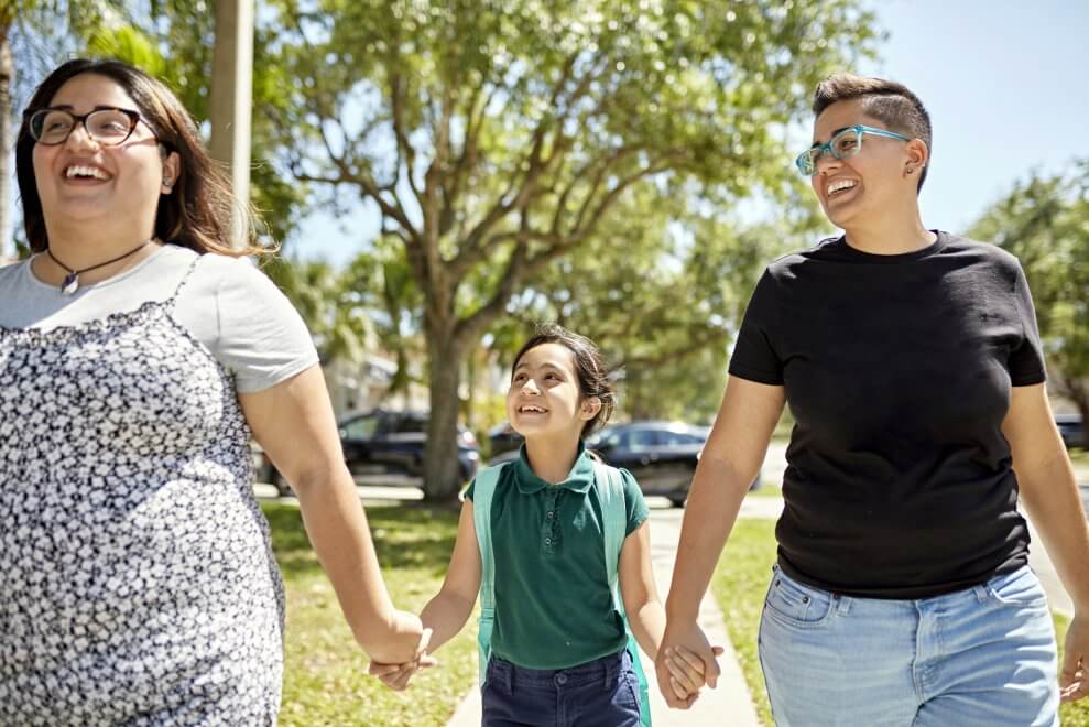 Two moms walk hand in hand with their daughter down a sidewalk on a sunny day.