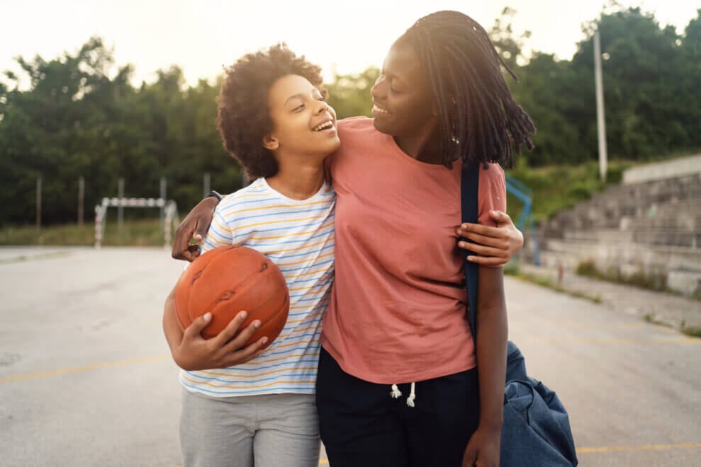 A mother and her daughter walk off an outdoor basketball court together, with their arms around each other. The daughter carries a basketball.