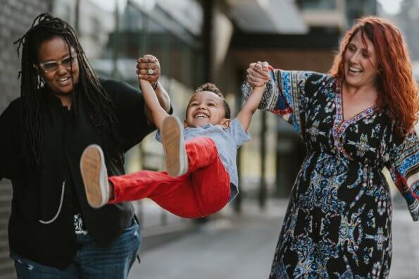 Two moms walk down the street, swinging their son by his arms. He has a big smile on his face.