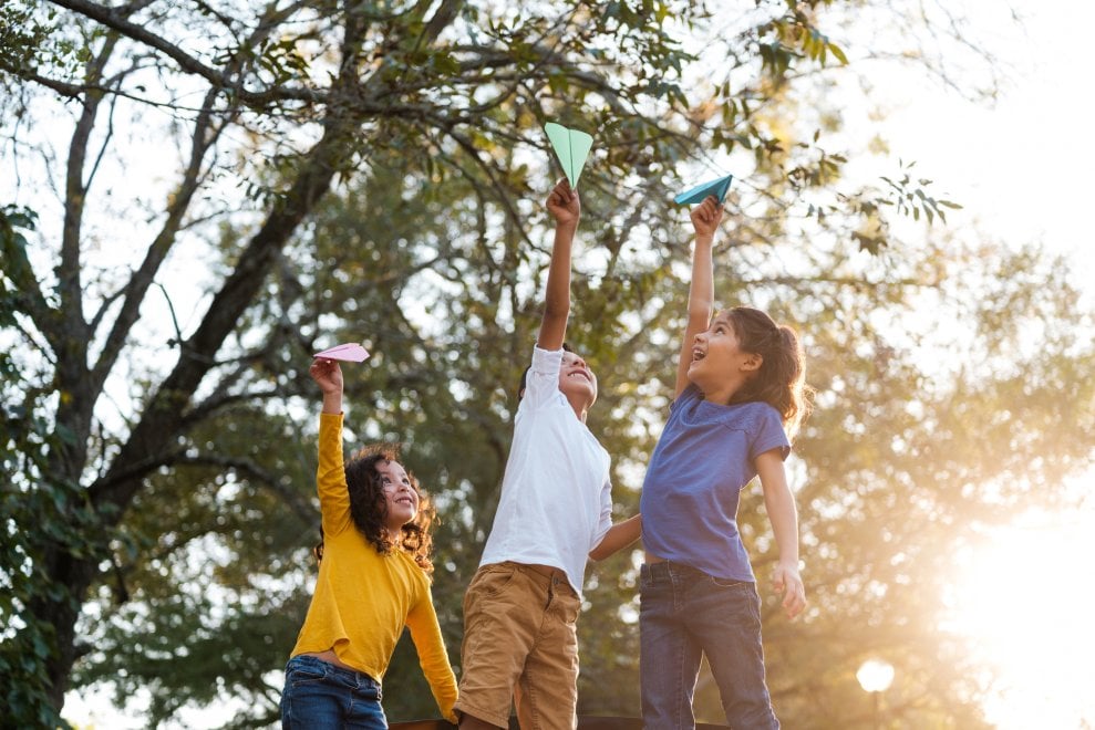 Three kids laugh and play with paper airplanes outside on a sunny day.