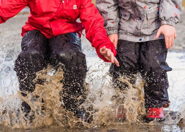 Two children in rain gear and rubber boots splash in a giant mud puddle.