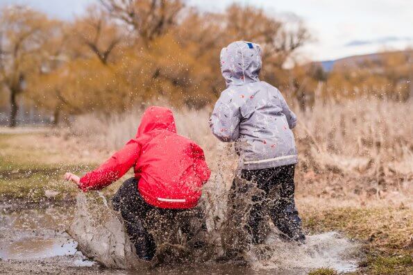 Two children in rain gear leap into a giant mud puddle, spraying the water into the air.