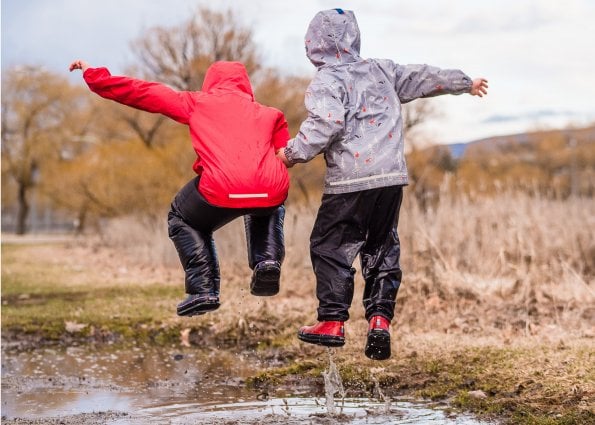 Two children in rain gear and rubber boots are about to jump into a giant mud puddle. They're in the air about to land in the water. 