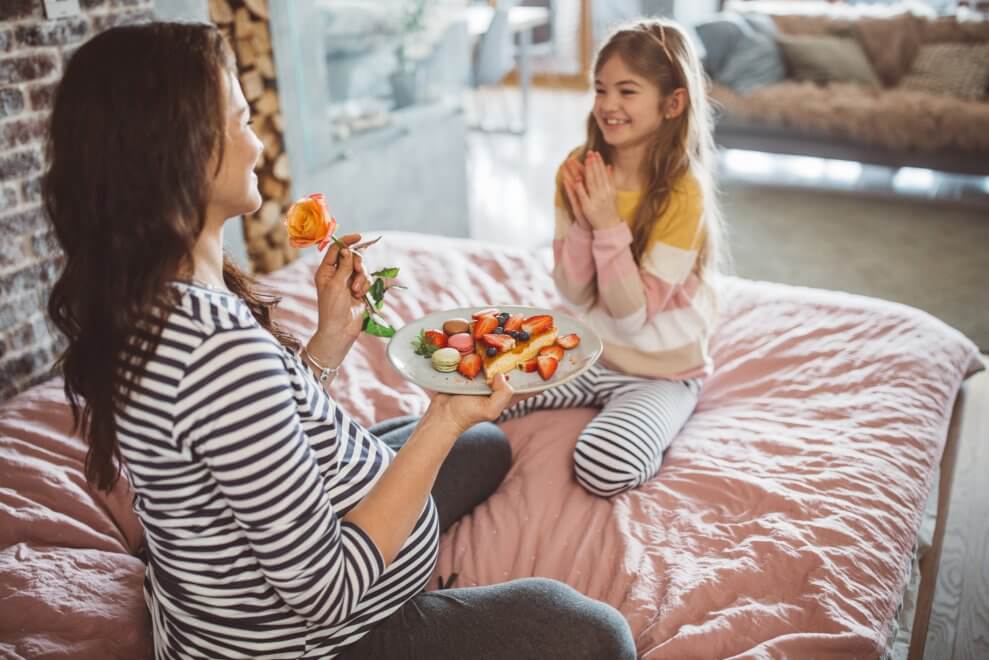A mother and her daughter sit on a bed together. The daughter has just handed her mom a plate of breakfast and a fresh flower.
