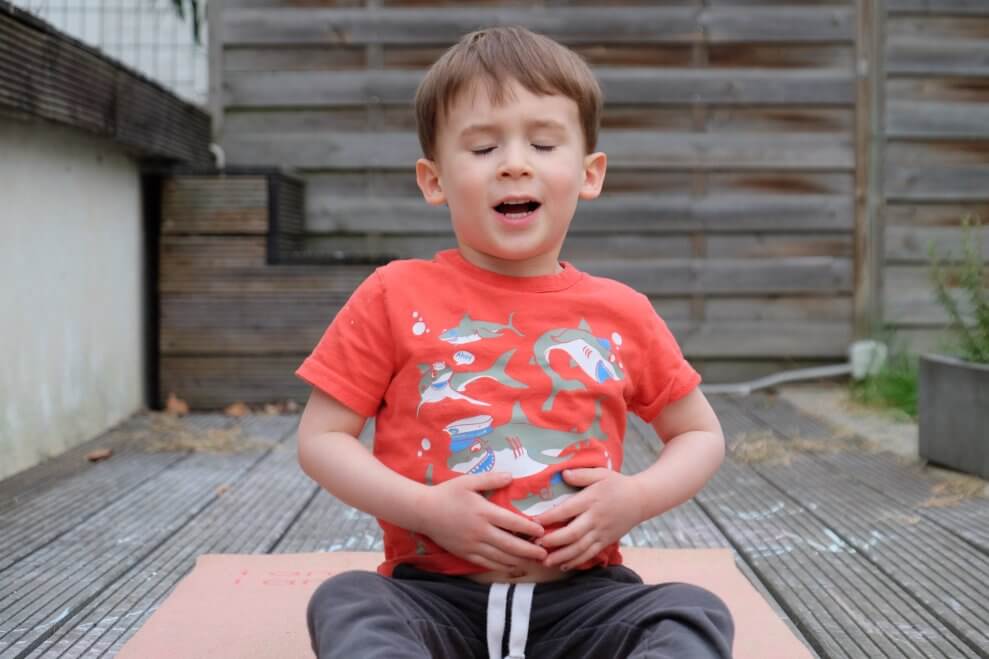 A toddler demonstrates belly breathing while sitting on a yoga mat.