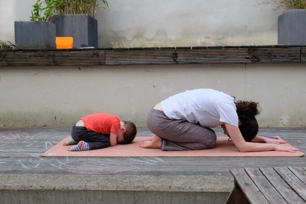 A mother and her toddler son do child's pose together on a yoga mat.