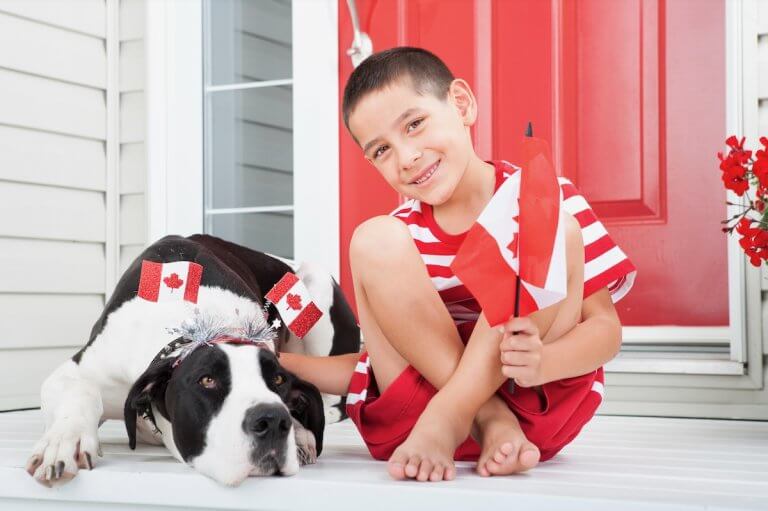 A boy sits on his front porch, wearing a red and white outfit. He holds mini Canada flags, and his dog lies beside him.