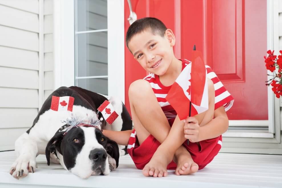 A boy sits on his front porch, wearing a red and white outfit. He holds mini Canada flags, and his dog lies beside him.