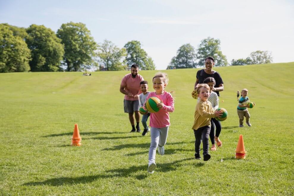 A group of young children runs around outside on a grassy field with two educators running behind them. Some of the children carry balls.