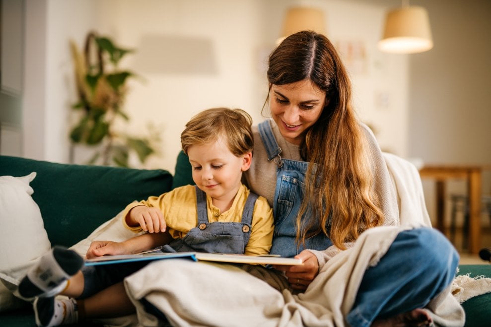 A mom and her son sit on the couch at home together and read a book. 