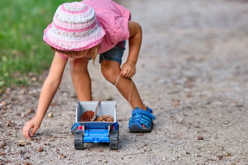 Little girl picks up stones and pinecones on the ground and puts them into a toy truck