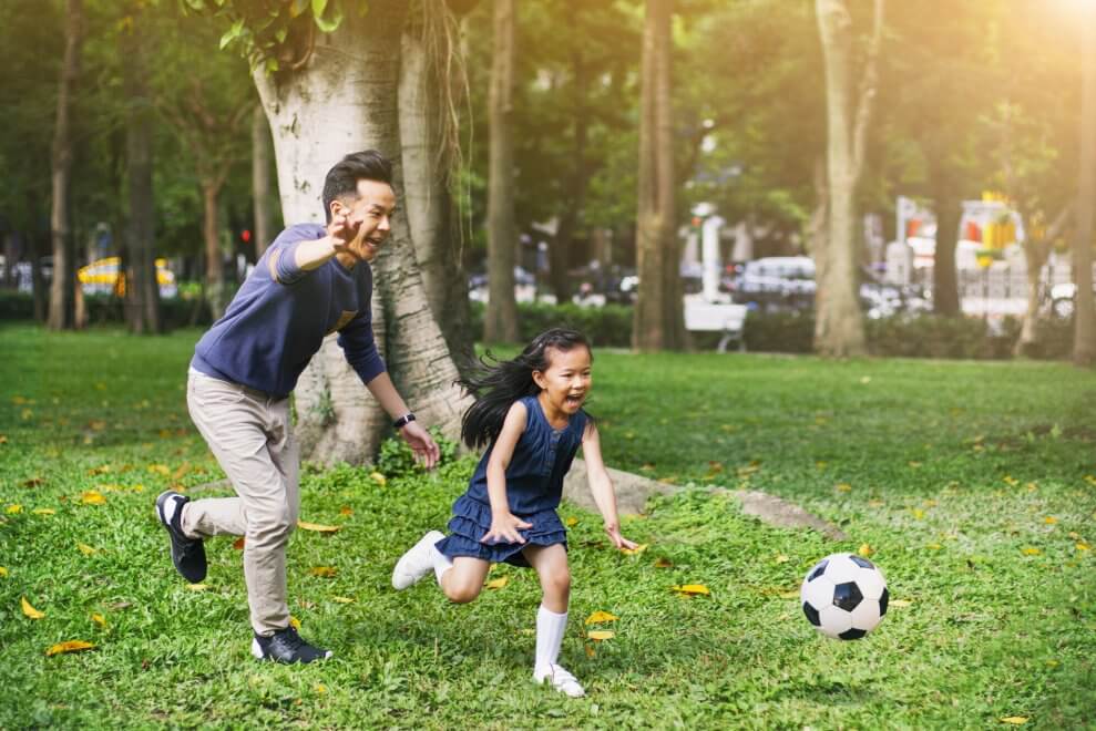 A girl and her father chase after a soccer ball, laughing