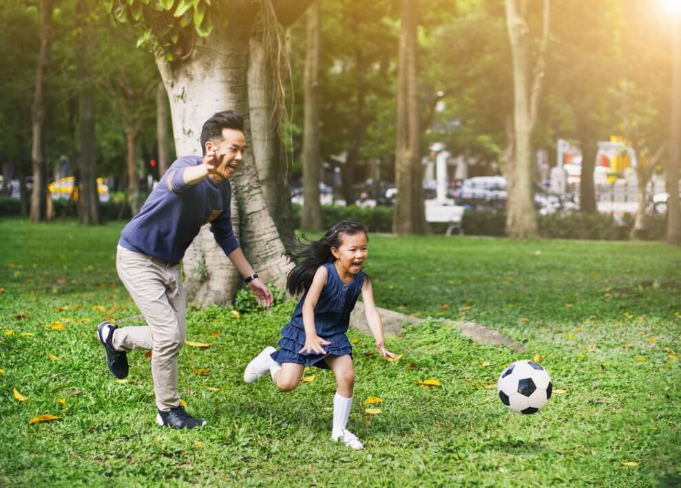 A girl and her father chase after a soccer ball, laughing