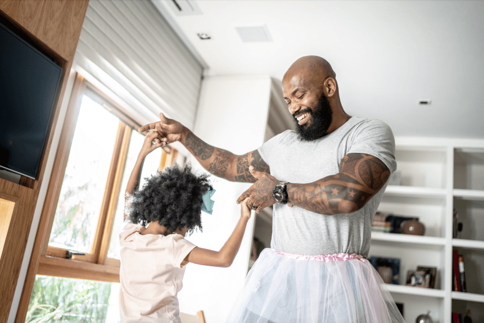 A dad and his young daughter dance together in their living room. The dad wears a pink tutu.
