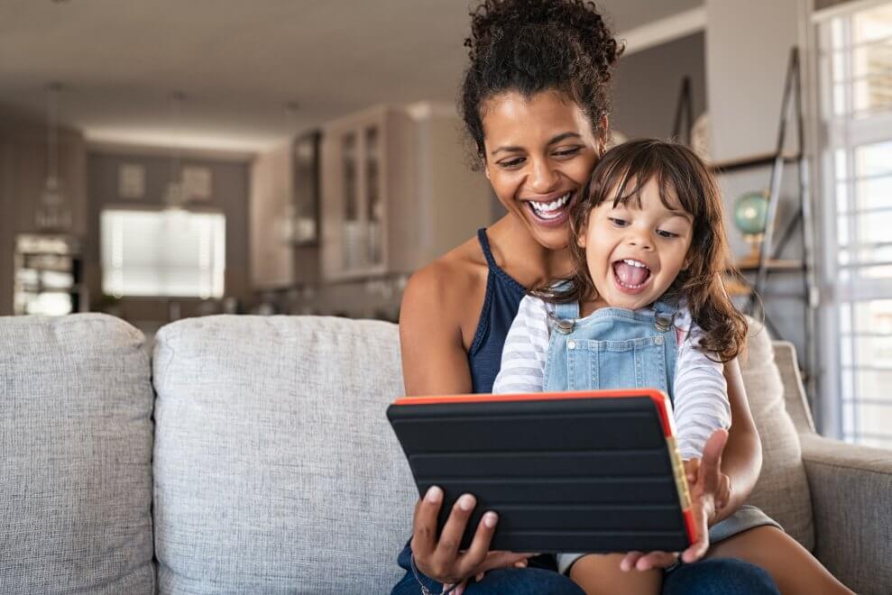 A mom and her young daughter use an iPad for a video call as they sit in their living room.