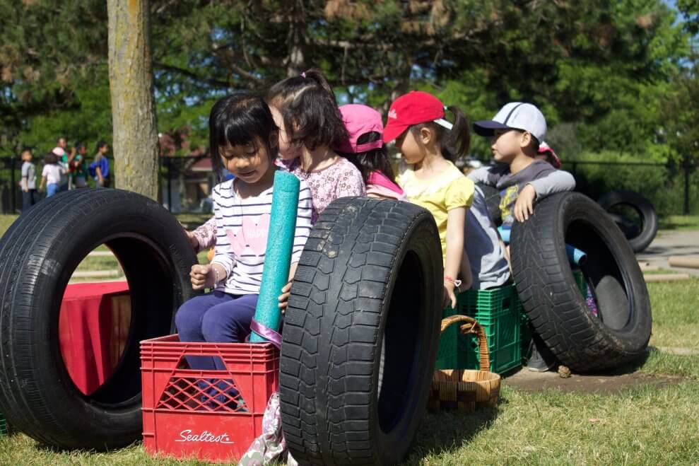 A group of children play outside in a grassy area with tires, pool noodles, and plastic crates.