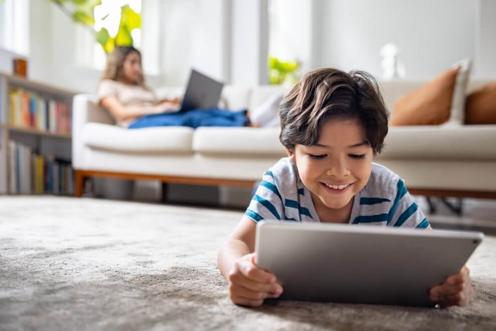 A son lies on his stomach on the living room floor and uses a tablet, while his mom uses a laptop on the couch behind him.