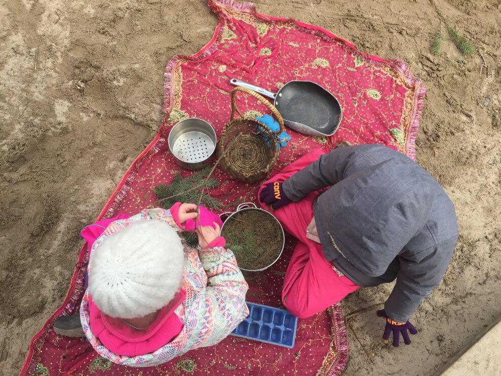 Two children sit on a blanket outside in the sand. They're playing with pots, pans, and tree boughs.