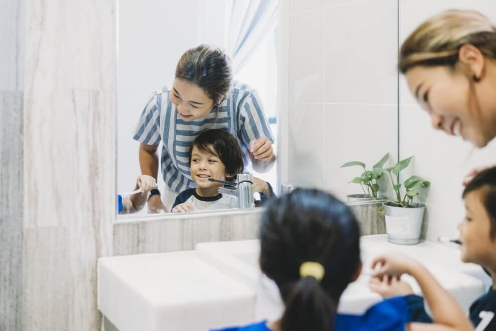 A mom stands with her two children at the bathroom sink while the kids brush their teeth.