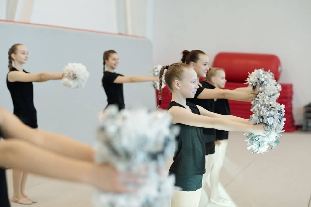 A group of girls do some basic cheer moves with pom poms at practice.