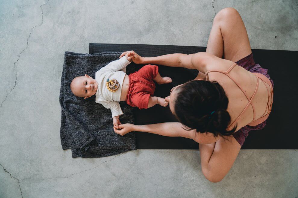 Mom sitting cross-legged while her baby lies in front of her on a yoga mat