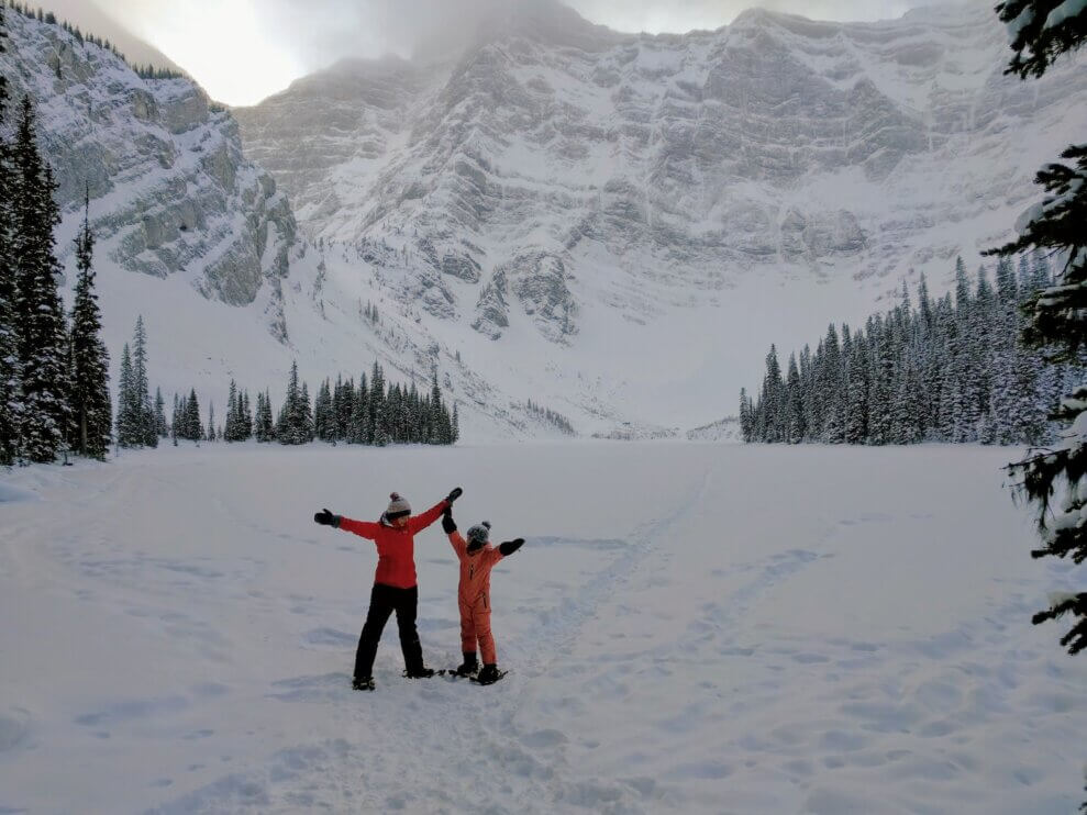 Mom and child stand in front of mountains, wearing snowshoes, with their arms raised to the sky.