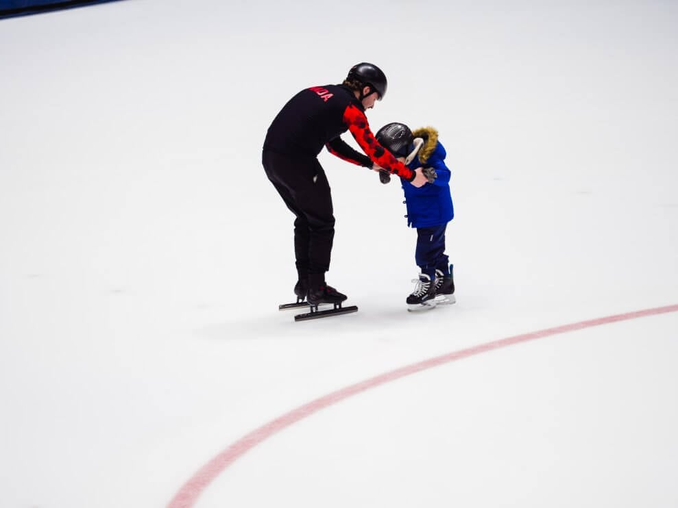 A speed skating coach helps a young child learn to skate by supporting their arms on the ice.