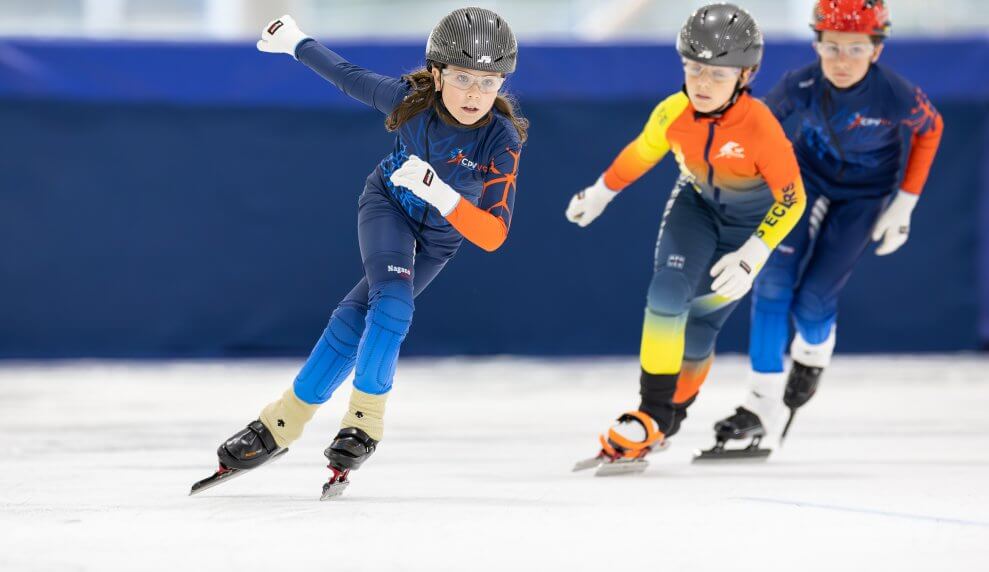 A young girl and two boys speed skate around a track. They're wearing skin suits, sport glasses, and helmets.