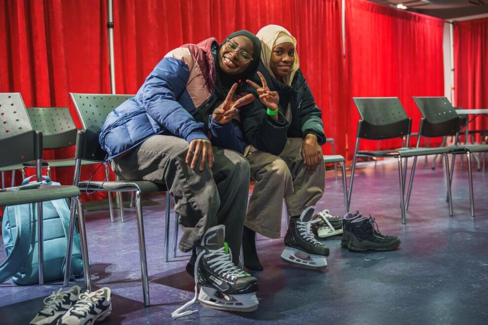 Two young women sit on chairs at an ice rink and put hockey skates on.