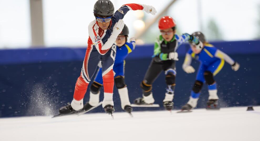 A group of children wearing skin suits, sport glasses, and helmets as they speed skate on a track.