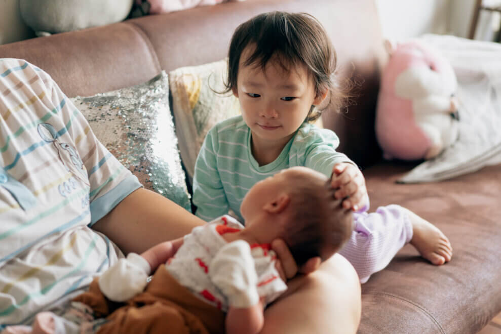 Parent sits on the couch, holding new baby, while toddler sibling sits beside the baby and touches its head