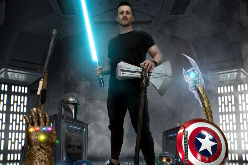 Glenn Higgins hold a lightsaber and stands with his superhero props around him