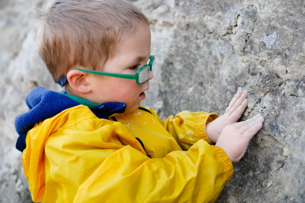 A young boy wearing glasses and a raincoat participates in sensory play by feeling a large boulder with his hands.