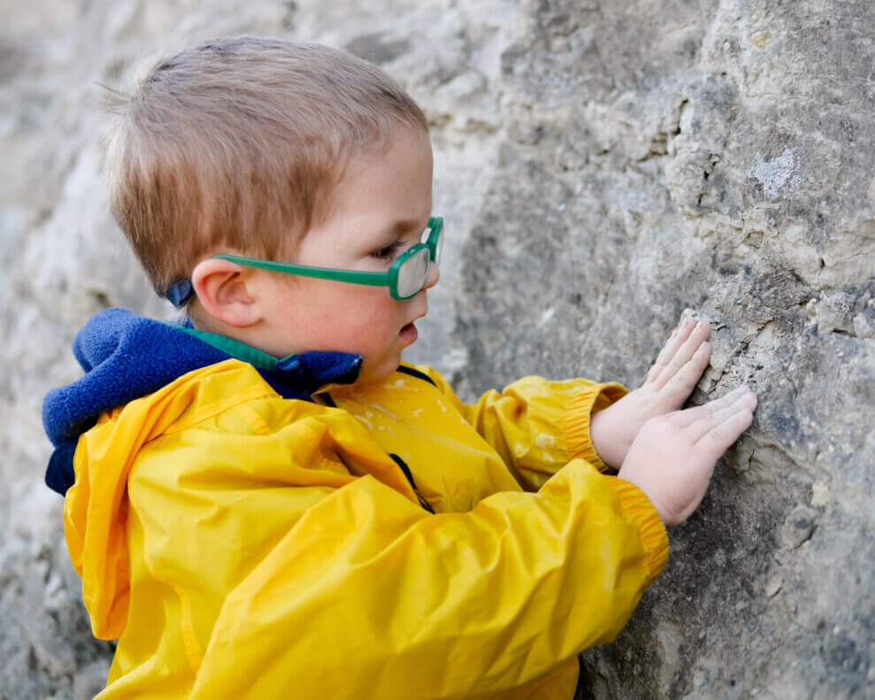Young boy wearing glasses touches a large rock wall 
