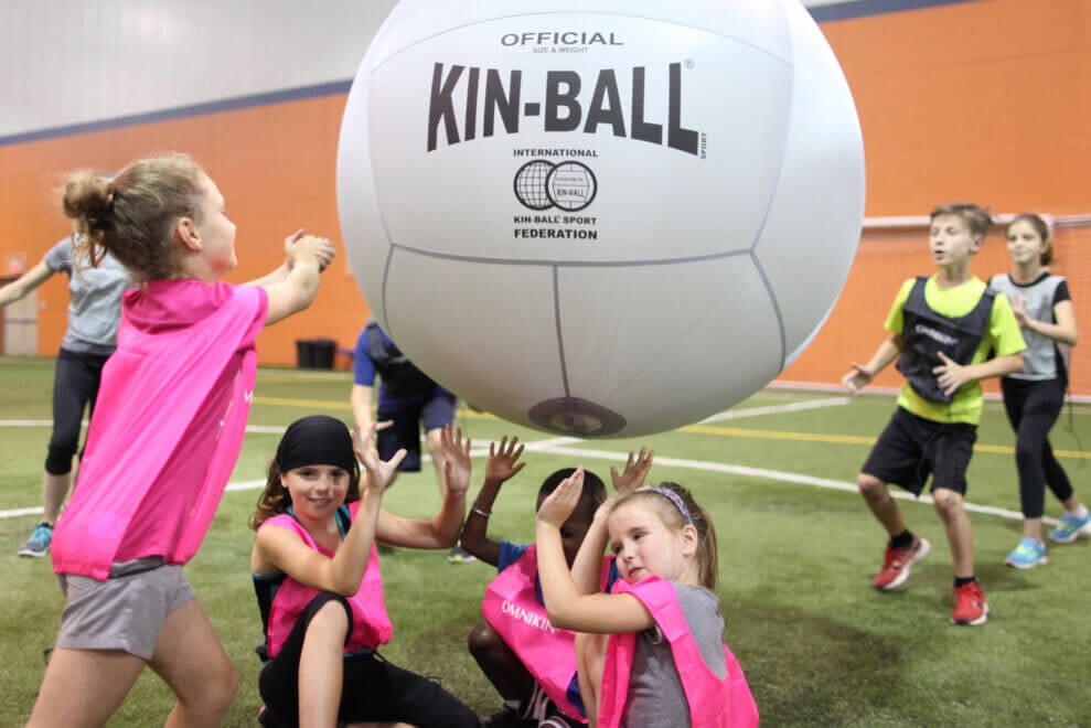 Group of children plays the sport of Kin-Ball indoors, with one girl about to hit the giant inflatable ball